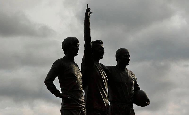 Foto van de standbeelden van George Best, Denis Law en Bobby Charlton bij Manchester United